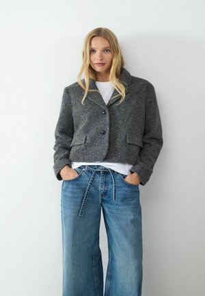 Grijze, kort gesneden wollen jas met een kraag en twee voorzakken, gecombineerd met lichtblauwe, wijde denim jeans en een wit t-shirt.