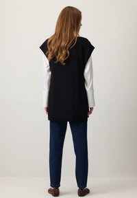 Hátulnézetből egy nő, hosszú hullámos hajjal, fekete ujjatlan pulóvert visel fehér blúz felett, kék farmert és leopárdmintás balerinacipőt.