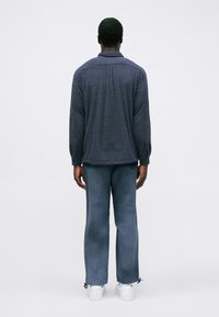 Μπλε σκούρο fleece μπουφάν με υφή, μακριά μανίκια και άνετη εφαρμογή, συνδυασμένο με ανοιχτό μπλε παντελόνι φαρδιάς γραμμής και λευκά αθλητικά παπούτσια.