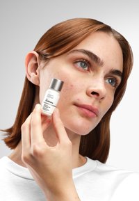 Witte fles met zilveren dop, gelabeld "The Ordinary." Model houdt het dicht bij haar gezicht, waarbij ze haar heldere huid en bruine haar laat zien.