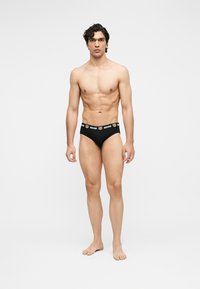 Modèle masculin debout pieds nus, portant un slip noir avec une ceinture de marque ornée de visages d'ours en peluche et du texte "MOSCHINO".