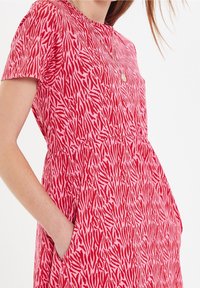 Punainen ja vaaleanpunainen kuviollinen mekko, jossa on lyhyet hihat, pyöreä kaula-aukko, vyötärösauma ja sivutaskut. Kevyt kangas, rento malli.