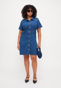 Blå denim skjortekjole med kortermede, knap foran og en figursyet silhuett. Kombinert med en mørk, flettet håndveske og spissede leiligheter.