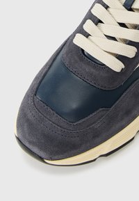 Közelebbi kép egy sötétkék sportcipőről, amely krémszínű fűzőkkel, velúr és bőr panelekből készült, valamint krémszínű gumitalppal rendelkezik.