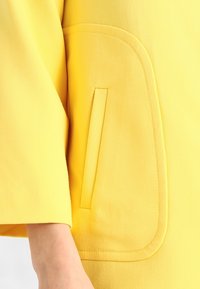 Gult ytterplagg med en texturerad, slät tyg. Har en stor, rund ficka och en något lös design med 3/4 ärmar.