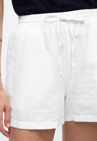 Valged linased lühikesed püksid kummipaela, nööriga detaili ja lõdva lõikega. Omaduseks on sile tekstuur ja serv, mis ulatub reie kohale.