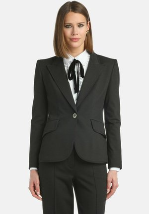 Blazer nero su misura con revers, chiusura a un bottone, due tasche frontali e una vestibilità strutturata sopra una camicia bianca con volant e nastro nero.