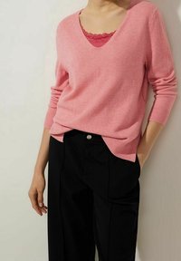 Rosa stickad tröja med V-ringning, ribbade ärmslut och sidofickor, lagd över en röd topp med spetsdetaljer, kombinerad med svarta löst sittande byxor.