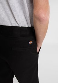 Black pants made of sturdy fabric; features a back pocket and a Dickies logo patch. Tapered design shown from the side with a gray shirt above.