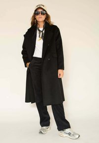 Longue veste noire sur une chemise blanche, associée à un pantalon noir et des baskets argentées. Accessoires : lunettes de soleil foncées et casquette beige.