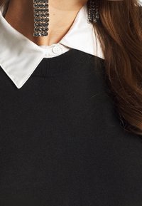 Donna con un maglione nero sopra una camicia bianca con colletto, orecchini a strass neri grandi, lunghi capelli castani visibili da un lato.