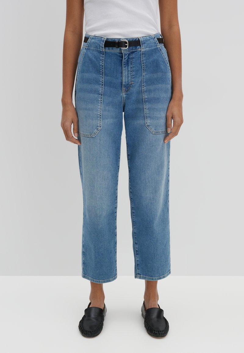 Jeans di denim azzurro chiaro a vita alta con gamba larga e tasche frontali, abbinati a scarpe nere slip-on. Texture liscia.