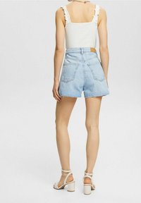 Shorts de mezclilla azul claro de cintura alta con dobladillo deshilachado, dos bolsillos traseros y una blusa sin mangas blanca con volante, combinados con sandalias de tacón grueso blancas.