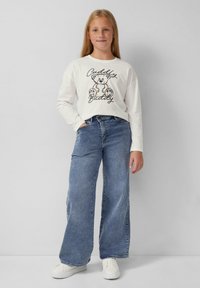 Langärmliges weißes Hemd mit schwarzem Bärenmotiv und Schrift, kombiniert mit hellblauen weiten Jeans und weißen Sneakers. Schlichter Hintergrund.