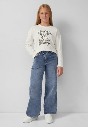 Langärmliges weißes Hemd mit schwarzem Bärenmotiv und Schrift, kombiniert mit hellblauen weiten Jeans und weißen Sneakers. Schlichter Hintergrund.