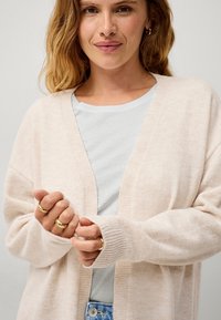 Cardigan aperto color beige chiaro realizzato in tessuto morbido con polsini a coste, indossato su una t-shirt azzurra chiara, con anelli d'oro sovrapposti sulle mani.