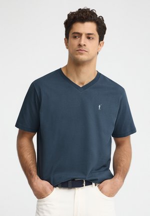 Man met donker krullend haar die een marineblauw V-hals T-shirt en crème broek draagt, staand met handen in de zakken tegen een witte achtergrond.