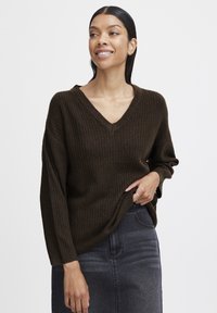 Suéter marrón de punto con escote en V y textura acanalada, con mangas largas y un corte relajado, combinado con una falda de mezclilla oscura.