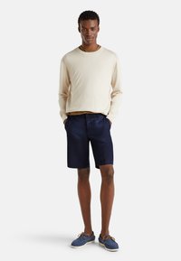 Béžová košile s dlouhým rukávem, námořnické šortky a modré boty s hnědými tkaničkami. Oblečení má uvolněný střih a jednoduchý design.