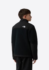 Crna flis jakna s kontrastnim gornjim dijelom od crnog najlona, s uspravnim ovratnikom i bijelim logotipom na leđima.