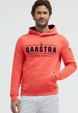 Mann trägt einen korallenfarbenen Kapuzenpullover mit dem Aufdruck „GAASTRA Nautical Supplies“ und Koordinaten, kombiniert mit weißen Hosen, stehend vor einem einfarbigen Hintergrund.