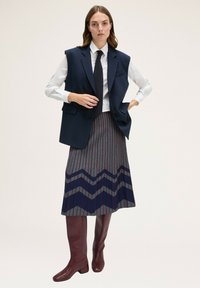 Marineblauer ärmelloser Blazer über einem weißen Shirt, kombiniert mit einem gemusterten grauen Rock, der marineblaue und burgunderfarbene Zickzacklinien aufweist, und kniehohen burgunderfarbenen Stiefeln.
