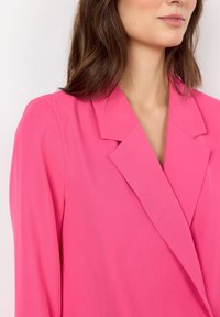 Fuksjowy żakiet z wyraźnym kołnierzem, długimi rękawami i gładką fakturą. Posiada kanciaste klapy i dopasowany krój.