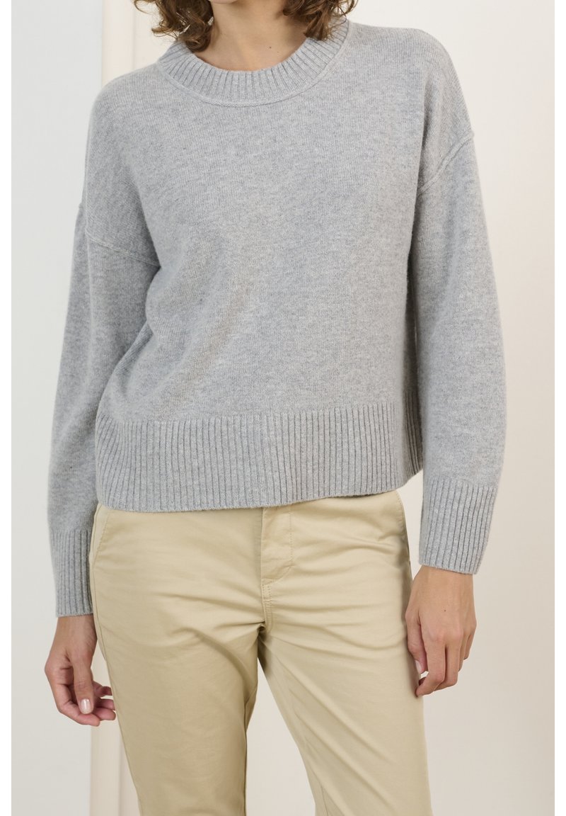 Femme portant un pull en maille côtelée gris clair et un pantalon beige, debout avec une main détendue le long du corps.