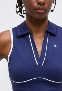 Γυναίκα φοράει navy μπλε αμάνικο τοπ με γιακά, άσπρες λεπτομέρειες και μικρό λογότυπο στο στήθος, σε απλό φωτεινό φόντο.