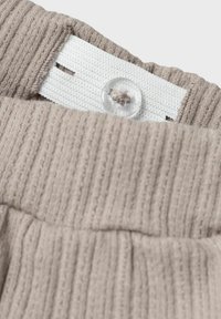 Beige geripptes Material mit elastischem Bund, ausgestattet mit einem weißen strukturierten Band und einem klaren Knopf oben, der einen sicheren Verschluss bietet.