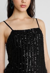 Svart paljettklänning med tunna spagettistremmar, med vertikala rader av paljetter för textur och glans mot en enkel tygbakgrund.