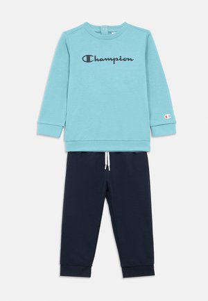 Hellblauer Pullover mit langen Ärmeln und einem schwarzen "Champion"-Logo, kombiniert mit marineblauen Jogginghosen mit Kordelzug und Bündchen.