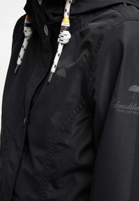 Chaqueta impermeable negra con capucha, que presenta cordones trenzados blancos, un logotipo impreso sutil y cierre de botón a presión. Textura mate suave.