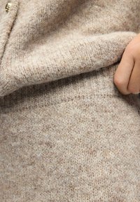 Étoffe de pull en mailles beige avec une finition texturée et un ourlet côtelé visible. Une main tient le tissu, mettant en avant sa douceur.