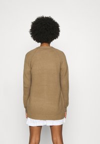 Person med lockigt hår som bär en texturerad brun långärmad tröja över en kort vit kjol, stående framför en enkel vit bakgrund.