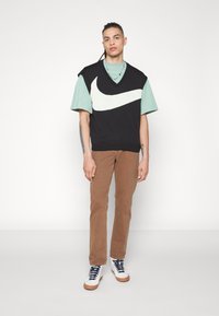 Schwarze ärmellose Strickjacke mit weißem Nike-Logo über einem mintgrünen T-Shirt, kombiniert mit braunen Hosen und weißen Sneakers mit blauen Akzenten.