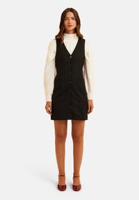 Abito nero a righe con scollatura a V, chiusura a bottoni e tasche laterali. Il modello lo abbina a un maglione bianco strutturato e tacchi rossi.