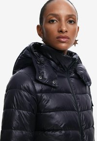 Giubbotto imbottito in un profondo blu navy, con zip frontale, cappuccio staccabile e una texture lucida a trapunte con cuciture visibili.