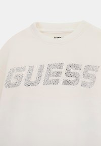 Krémová mikina s veľkým strieborným logom "GUESS" ozdobeným kamienkami na prednej strane. Mäkká tkanina s dizajnom okrúhleho výstrihu.