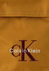 Marka Calvin Klein na teksturowanej musztardowej tkaninie, z haftowanym napisem "CK" w ciemnym bordowym kolorze oraz napisem "Calvin Klein" poniżej w białym kolorze.