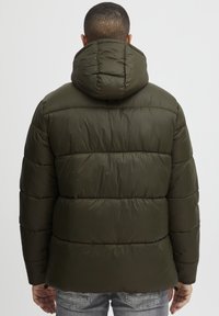Puffjacka i mörk olivgrön med vaddering, en dragsko i huvan och elastiska ärmslut. Har en slät, glänsande yta.
