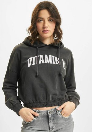 Sudadera corta negra con cordones; presenta el texto "VITAMIN D" en blanco. Textura suave, corte relajado y dobladillo elástico. Combinada con jeans grises.