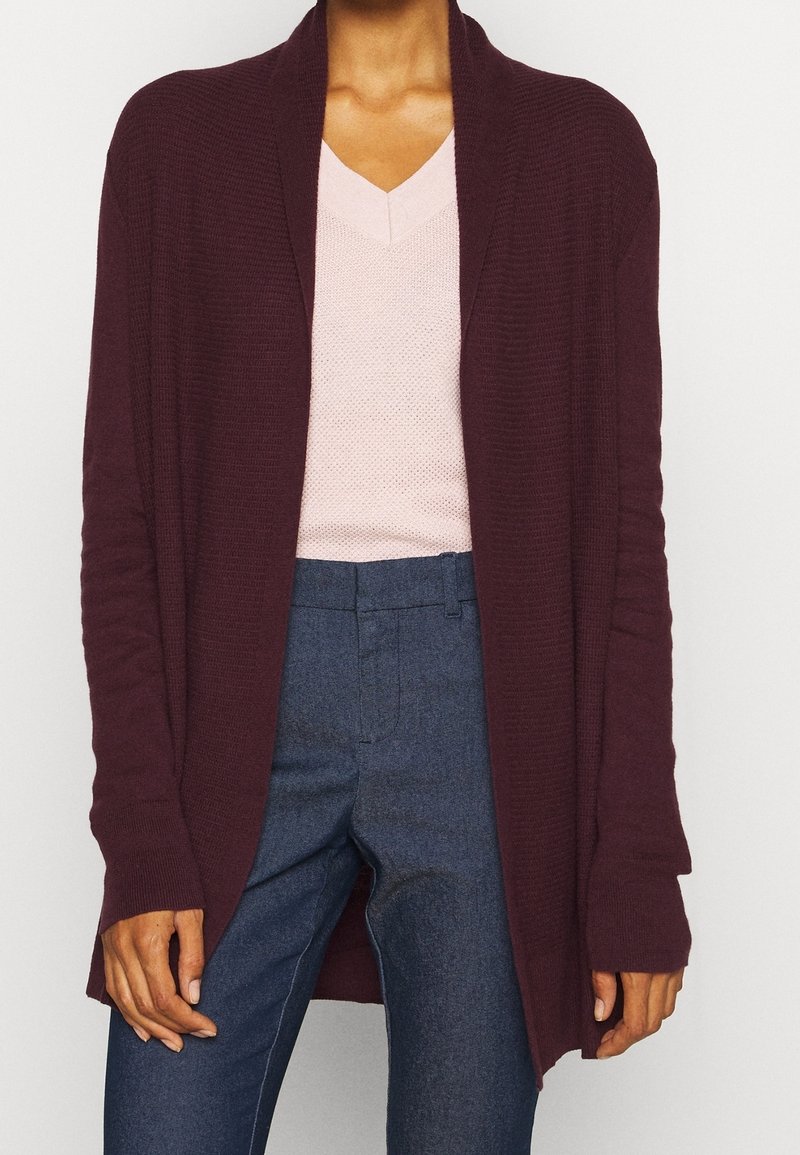Femme portant un cardigan bordeaux foncé ouvert sur le devant, un pull rose clair à col en V et un pantalon bleu taille haute sur un fond uni.