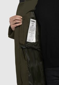 Olivenfarvet jakke med en glat yderskal, indvendig foring, elastisk snor i taljen og et plejelabel synligt inde i lommen.