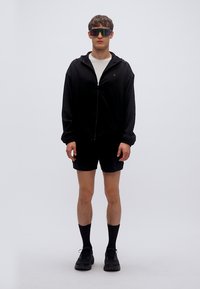 Μαύρο ελαφρύ τζάκετ με φερμουάρ, λευκό μπλουζάκι, μαύρες βερμούδες, καλτσάκια μέχρι το γόνατο και χοντρά μαύρα αθλητικά παπούτσια. Ανακλαστικά γυαλιά ηλίου με έντονο σχέδιο.