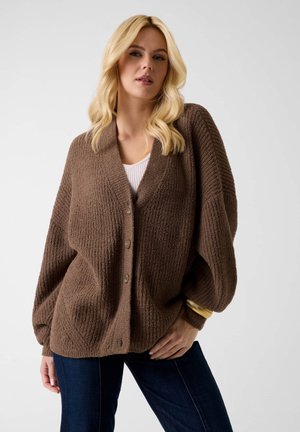 Blonde vrouw met een bruine, getextureerde, geknoopte cardigan over een witte top en donkere spijkerbroek, poserend met één hand op haar heup.