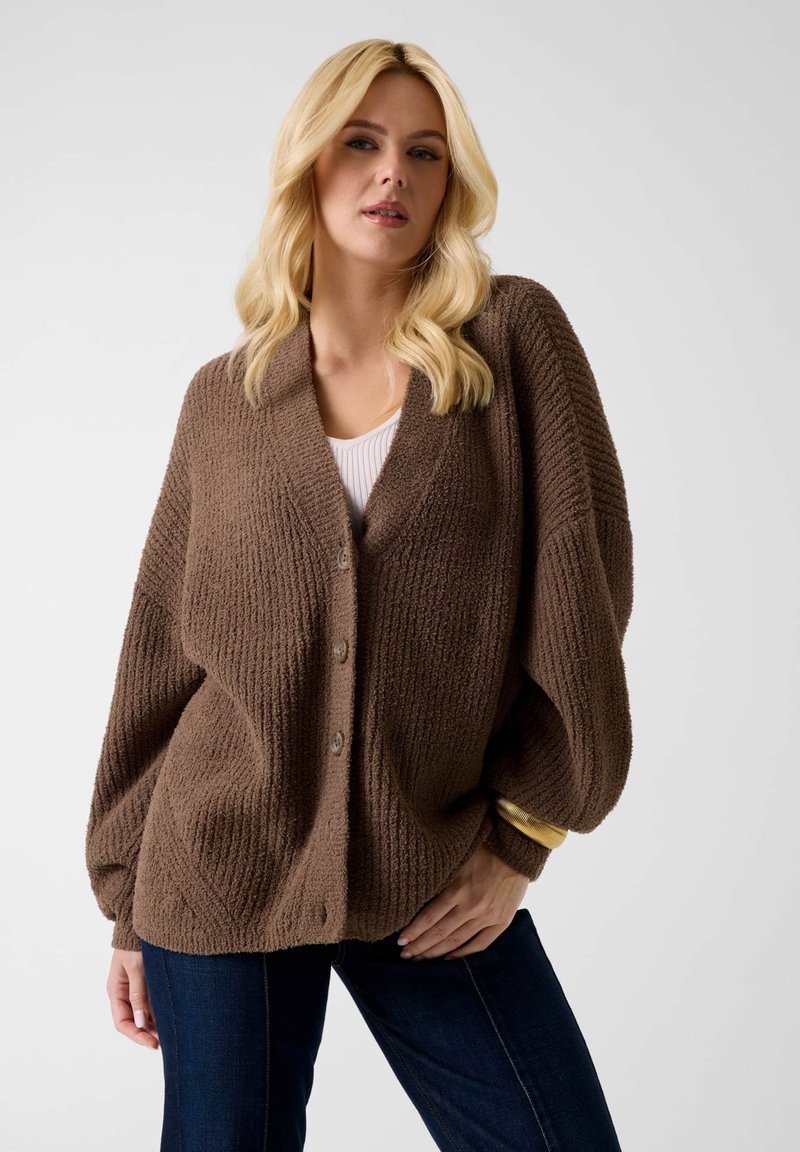 Plavuša koja nosi smeđi teksturirani kardigan s gumbima preko bijele majice i tamnih traperica, pozirajući s jednom rukom na bokovima.