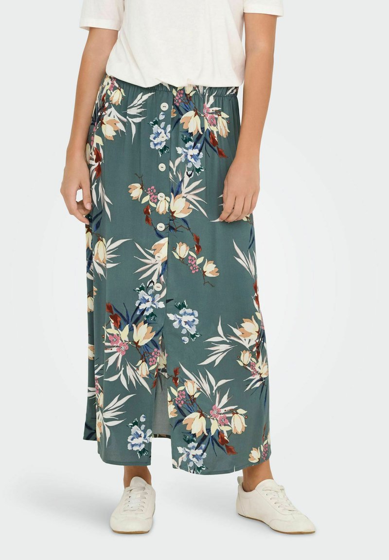 Lang, grøn blomsterkjole med knaplukning foran, elastisk talje og slidser i siden. Båret med hvide sneakers.
