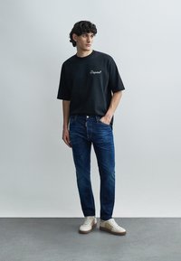Schwarzes kurzärmeliges T-Shirt mit gesticktem Logo, kombiniert mit blauen Slim-Fit-Jeans und beigen Sneakers mit strukturierten Akzenten.