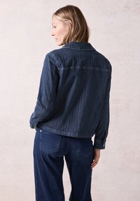 Tummansinisestä denim-takista, jossa on valkoiset raidoitukset, löytyy kauluskohouma ja pitkät hihat. Takki on yhdistetty yhdisteleviin tummiin housuihin.